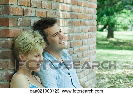 Couple sitting outdoors, leaning against brick wall, smiling View Large Photo Image Stock Image - Couple sitting outdoors, leaning against brick wall, smiling. Fotosearch