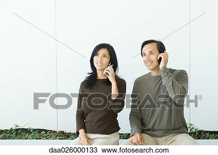Couple sitting side by side, both using cell phones, smiling View Large Photo Image Stock Image - Couple sitting side by side, both using cell phones, smiling. Fotosearch