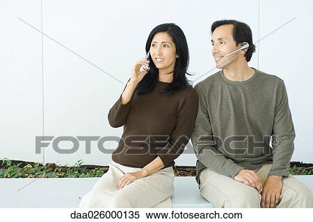 Couple sitting side by side, woman using cell phone, man wearing headset, both smiling View Large Photo Image Stock Photography - Couple sitting side by side, woman using cell phone, man wearing headset, both smiling. Fotosearch