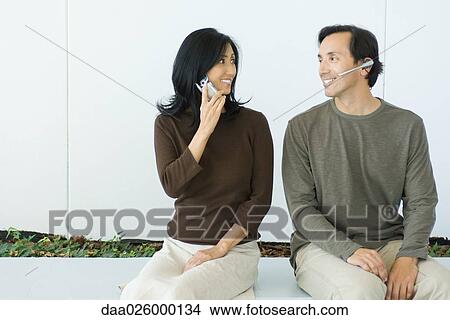 Couple sitting, smiling at each other, woman using cell phone, man wearing headset View Large Photo Image Picture - Couple sitting, smiling at each other, woman using cell phone, man wearing headset. Fotosearch