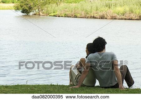 Stock Photography - Couple sitting together by lake, rear view. Fotosearch