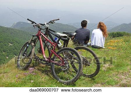 Couple sitting together on hill, looking at view, bicycles parked behind them View Large Photo Image Stock Photo - Couple sitting together on hill, looking at view, bicycles parked behind them. Fotosearch