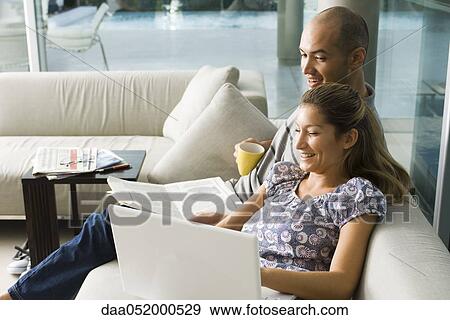 Couple sitting together on sofa, laughing at laptop computer View Large Photo Image Stock Photo - Couple sitting together on sofa, laughing at laptop computer. Fotosearch
