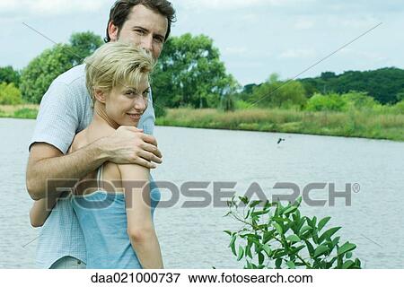 Couple standing by lake, looking over shoulders at camera View Large Photo Image Stock Photo - Couple standing by lake, looking over shoulders at camera. Fotosearch