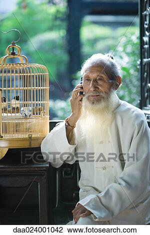 Stock Image - Elderly man wearing traditional Chinese clothing, using cell phone, next to bird cage. Fotosearch