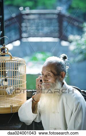 Stock Image - Elderly man wearing traditional Chinese clothing, using cell phone, next to bird cage. Fotosearch