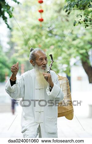 Stock Image - Elderly man wearing traditional Chinese clothing carrying bird cage over shoulder, holding up thumb. Fotosearch