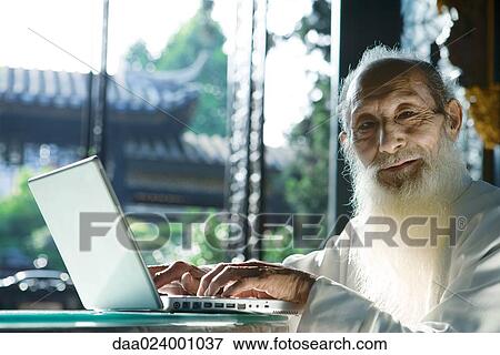 Stock Photo - Elderly man wearing traditional Chinese clothing using laptop, smiling at camera. Fotosearch