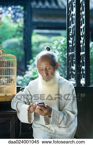 Stock Photography - Elderly man wearing traditional Chinese clothing, using cell phone, next to bird cage. Fotosearch