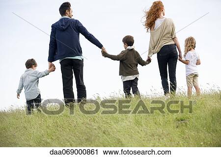 Stock Image - Family walking hand-in-hand in field, rear view. Fotosearch