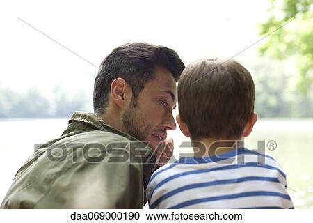 Stock Image - Father and son sitting beside lake, talking, rear view. Fotosearch