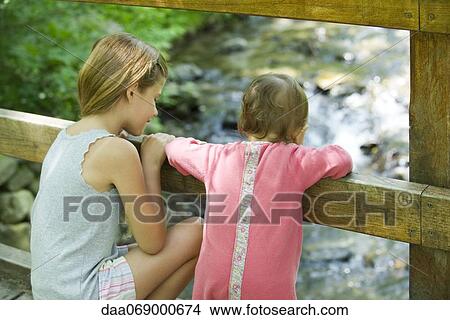 Picture - Girl and baby sister looking over railing of bridge at stream below. Fotosearch
