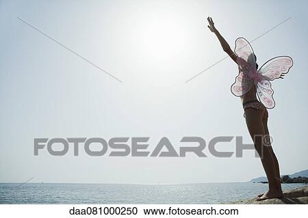Stock Image - Girl in bikini wearing butterfly costume, standing on edge of cliff with arms raised. Fotosearch