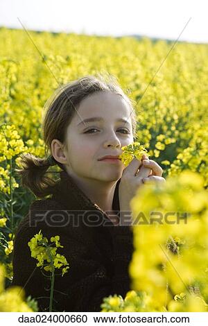 Stock Image - Girl in field of yellow flowers, smelling blossom, looking at camera. Fotosearch