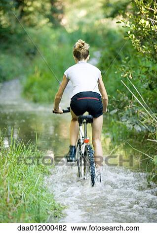 Stock Image - Girl riding through water on bike, rear view. Fotosearch