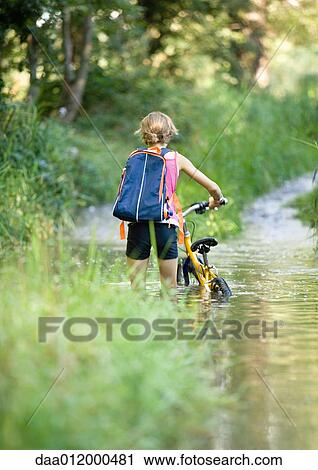 Stock Image - Girl walking through water with bike, rear view. Fotosearch