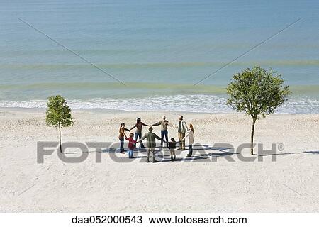 Stock Image - Group holding hands in circle on beach between two trees. Fotosearch