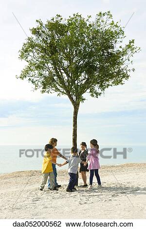 Stock Image - Group of children holding hands in circle around solitary tree on beach. Fotosearch