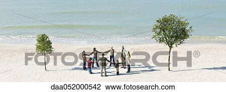 Stock Image - Group of people holding hands in circle between two trees on beach. Fotosearch