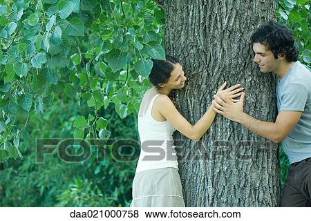 Man and woman leaning against either side of tree trunk, looking at each other, man holding woman's hand View Large Photo Image Stock Photo - Man and woman leaning against either side of tree trunk, looking at each other, man holding woman's hand. Fotosearch