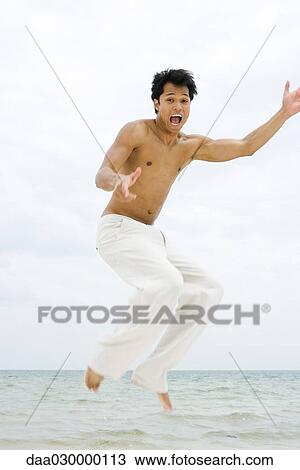 Stock Image - Man at the beach, jumping in the air, looking at camera, mouth open. Fotosearch