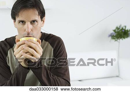 Stock Photo - Man holding cup with both hands, blowing into cup, looking at camera. Fotosearch