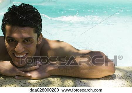 Stock Image - Man holding on to edge of swimming pool, head resting on arms, smiling at camera. Fotosearch