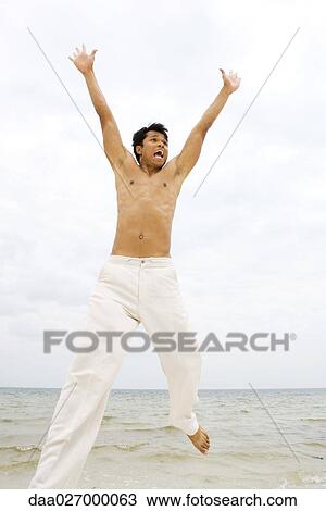 Stock Image - Man jumping in the air at the beach, arms raised, looking away. Fotosearch