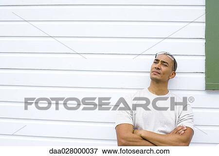 Stock Photo - Man leaning against side of house, arms folded, looking up. Fotosearch