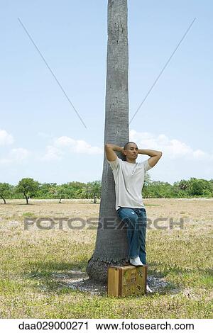 Stock Image - Man leaning against tree trunk, hands behind head, one foot resting on suitcase. Fotosearch