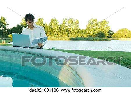 Stock Photograph - Man leaning on edge of pool, studying blueprints and using laptop. Fotosearch