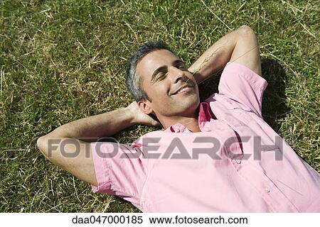 Stock Photography - Man lying on grass with hands behind head and eyes closed, sprig of grass in mouth. Fotosearch