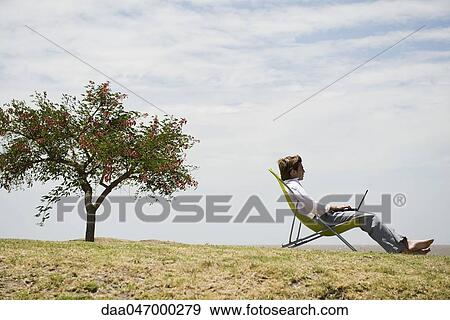 Stock Photo - Man relaxing in lounge chair outdoors, using laptop computer. Fotosearch