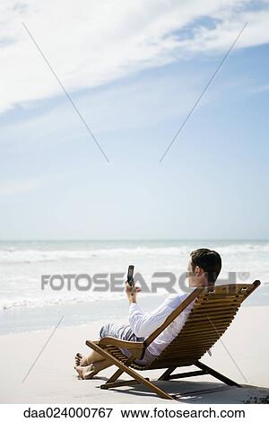 Man sitting in chair on beach, using cell phone, side view View Large Photo Image Stock Photo - Man sitting in chair on beach, using cell phone, side view. Fotosearch