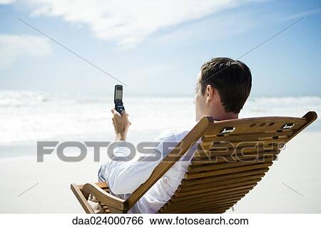 Stock Photograph - Man sitting in chair on beach, using cell phone, rear view. Fotosearch