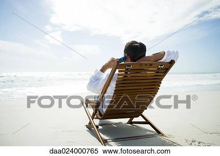 Stock Photography - Man sitting in chair on beach, using cell phone, rear view. Fotosearch