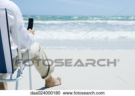 Man sitting in folding chair on beach, holding cell phone, cropped, rear view View Large Photo Image Stock Image - Man sitting in folding chair on beach, holding cell phone, cropped, rear view. Fotosearch