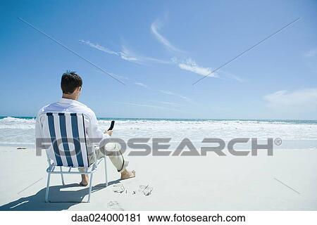 Man sitting in folding chair on beach, holding cell phone, rear view View Large Photo Image Stock Image - Man sitting in folding chair on beach, holding cell phone, rear view. Fotosearch