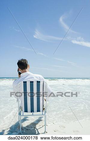 Man sitting in folding chair on beach, using cell phone, rear view View Large Photo Image Stock Photo - Man sitting in folding chair on beach, using cell phone, rear view. Fotosearch
