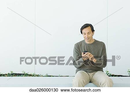 Man sitting, looking down at cell phone, smiling, front view View Large Photo Image Stock Image - Man sitting, looking down at cell phone, smiling, front view. Fotosearch