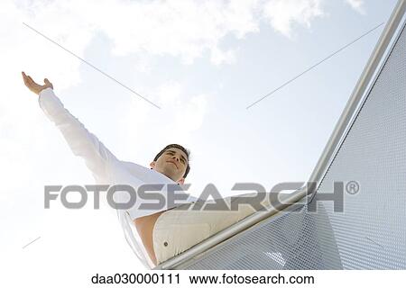 Stock Image - Man sitting on balcony, one arm raised, low angle view. Fotosearch