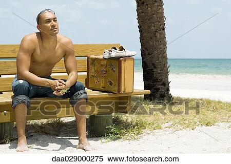 Stock Photography - Man sitting on bench at the beach, suitcase beside him, looking away. Fotosearch