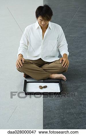 Stock Photo - Man sitting on floor, looking down at rock garden. Fotosearch