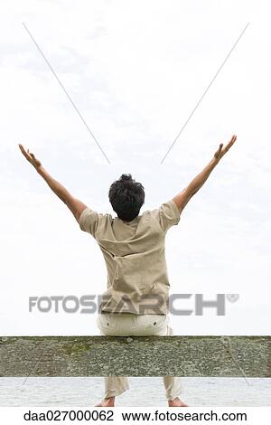 Stock Image - Man sitting on guard rail with arms raised, rear view. Fotosearch