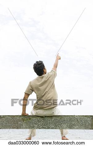 Man sitting on rail and pointing up, facing ocean, rear view View Large Photo Image Stock Image - Man sitting on rail and pointing up, facing ocean, rear view. Fotosearch