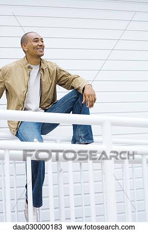 Man sitting on railing, looking away, smiling View Large Photo Image Stock Image - Man sitting on railing, looking away, smiling. Fotosearch