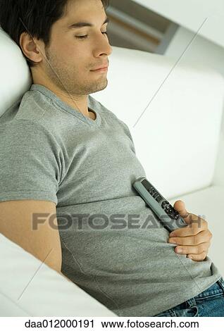 Man sitting on sofa, holding remote control against chest View Large Photo Image Stock Image - Man sitting on sofa, holding remote control against chest. Fotosearch