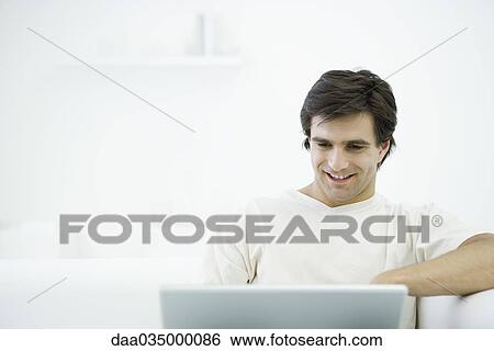 Man sitting on sofa, using laptop View Large Photo Image Stock Photograph - Man sitting on sofa, using laptop. Fotosearch