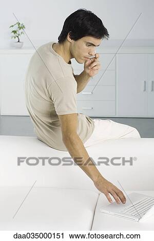 Man sitting on top of couch, using laptop and cell phone View Large Photo Image Stock Image - Man sitting on top of couch, using laptop and cell phone. Fotosearch