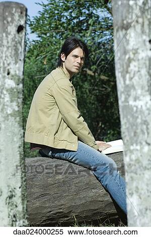 Stock Photography - Man sitting on tree trunk with book, looking away. Fotosearch